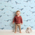 Lovely child standing in front of Whimsy Tots Whale Cloud Voyage Wallpaper in Sky Blue with whale, cloud, splash in design.