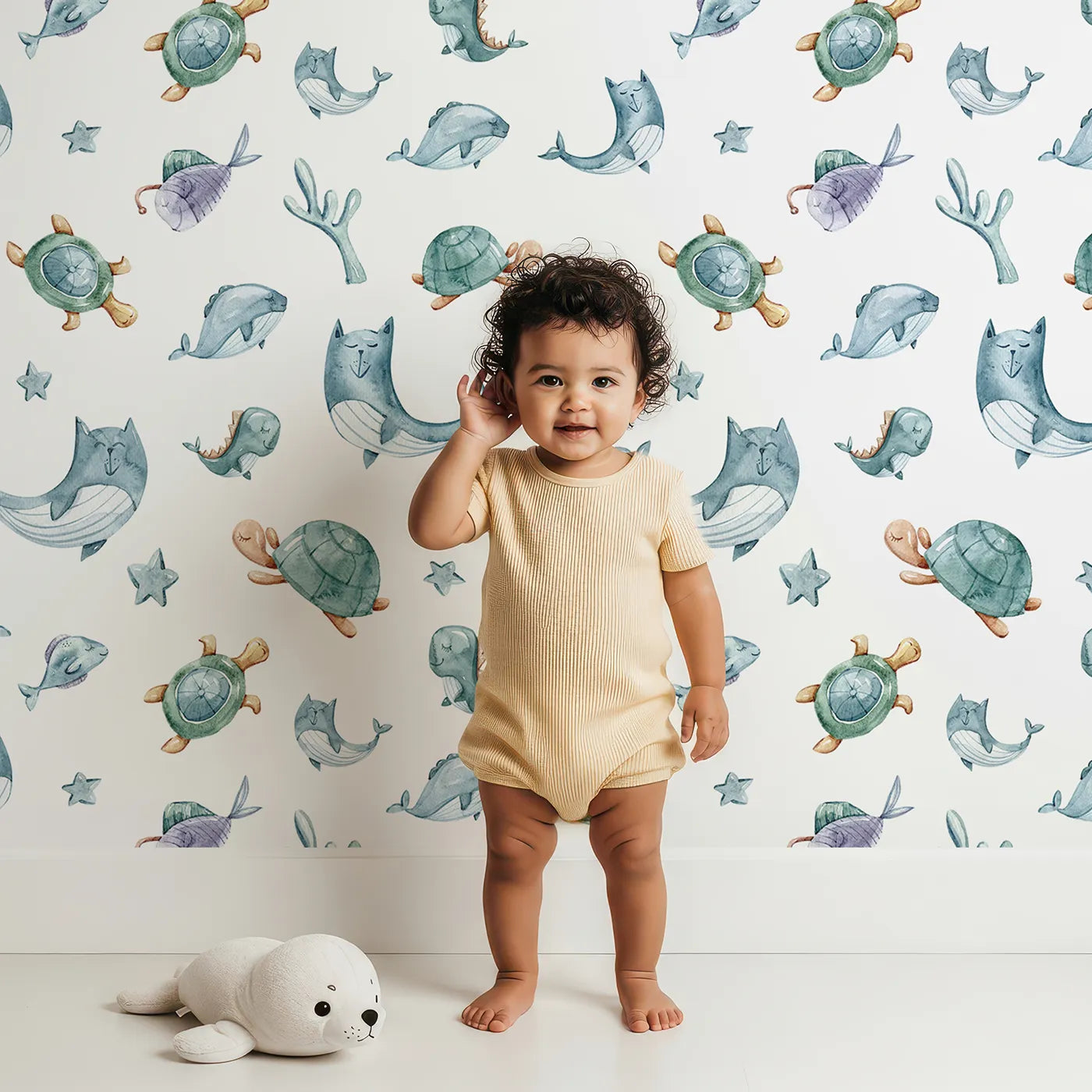 Lovely child standing in front of Whimsy Tots Gentle Sea Friends Wallpaper in White with whale, turtle, dolphin, fish, coral, seashell, seaweed, star in design.