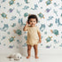 Lovely child standing in front of Whimsy Tots Gentle Sea Friends Wallpaper in White with whale, turtle, dolphin, fish, coral, seashell, seaweed, star in design.