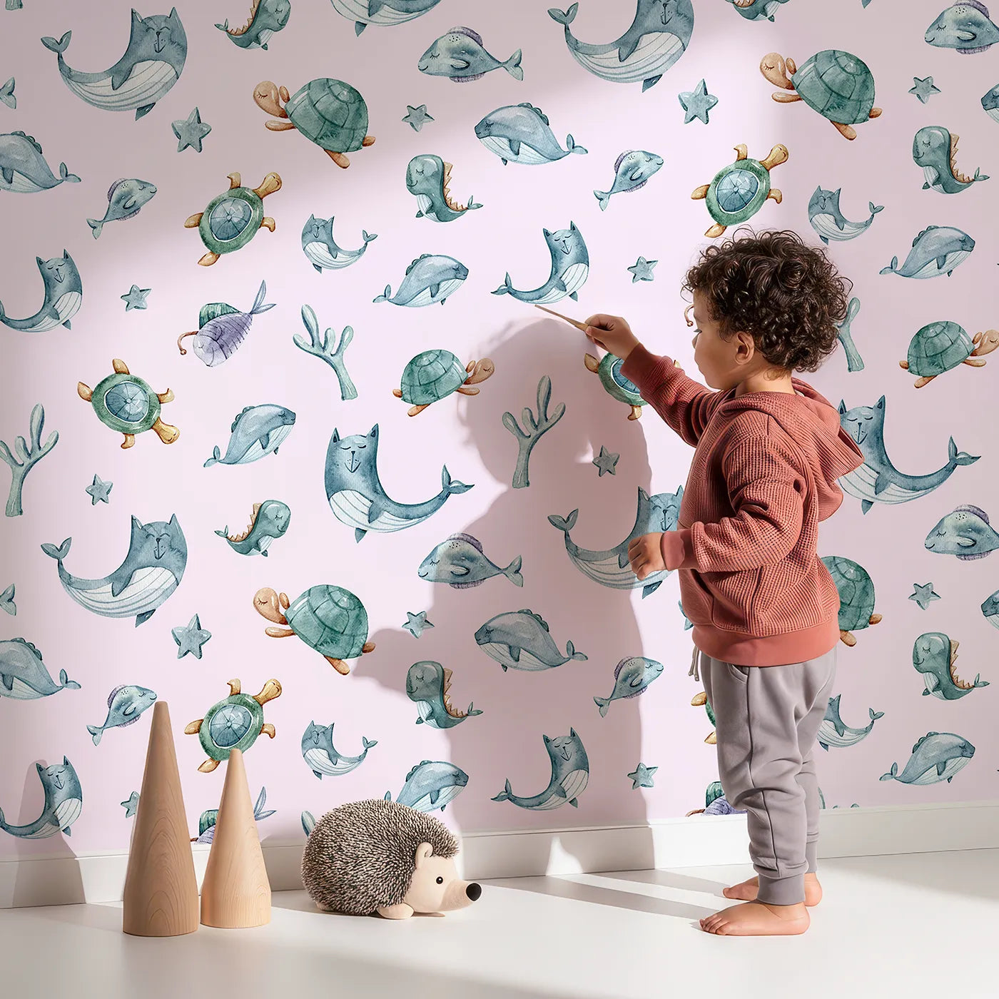 Lovely child standing in front of Whimsy Tots Gentle Sea Friends Wallpaper in Pink with whale, turtle, dolphin, fish, coral, seashell, seaweed, star in design.