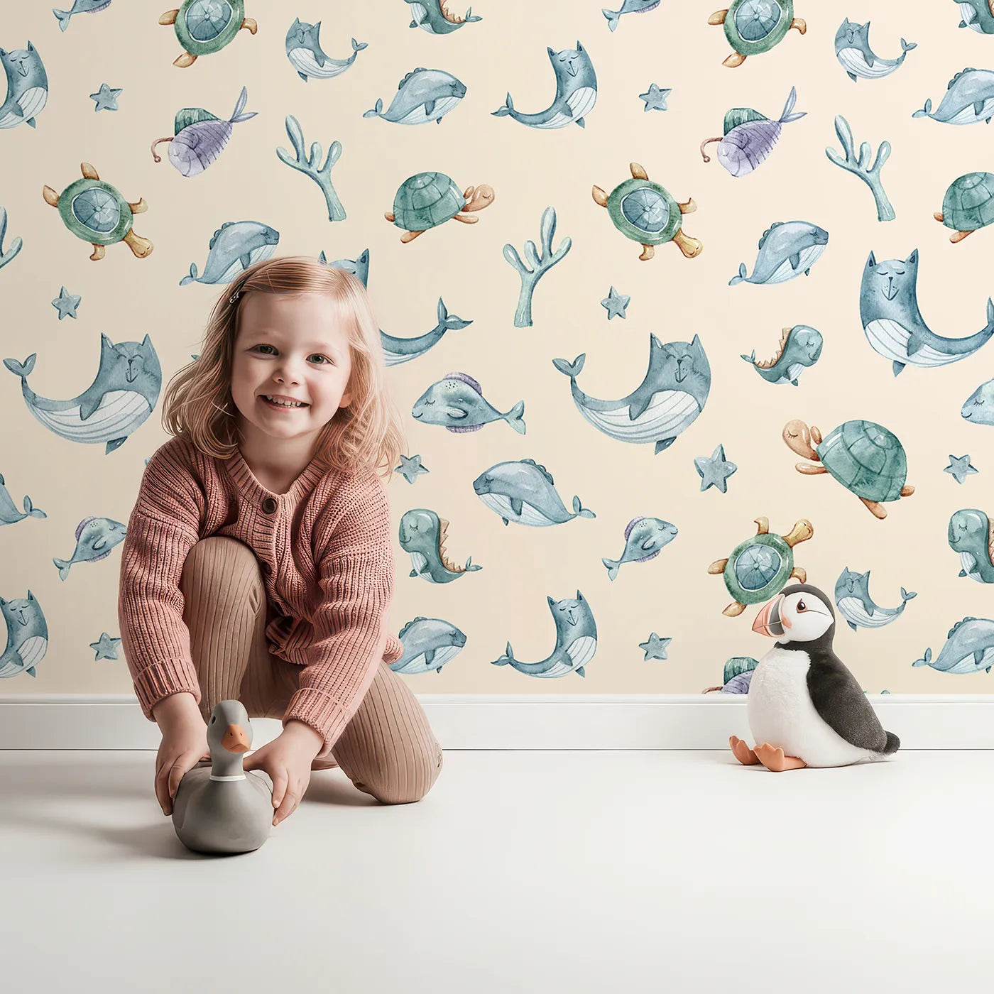 Lovely child standing in front of Whimsy Tots Gentle Sea Friends Wallpaper in Peach with whale, turtle, dolphin, fish, coral, seashell, seaweed, star in design.