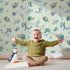 Lovely child standing in front of Whimsy Tots Gentle Sea Friends Wallpaper in Mint Green with whale, turtle, dolphin, fish, coral, seashell, seaweed, star in design.