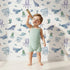 Lovely child standing in front of Whimsy Tots Gentle Ocean Parade Wallpaper in White with whale, narwhal, octopus, jellyfish, fish in design.
