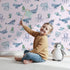 Lovely child standing in front of Whimsy Tots Gentle Ocean Parade Wallpaper in Pink with whale, narwhal, octopus, jellyfish, fish in design.