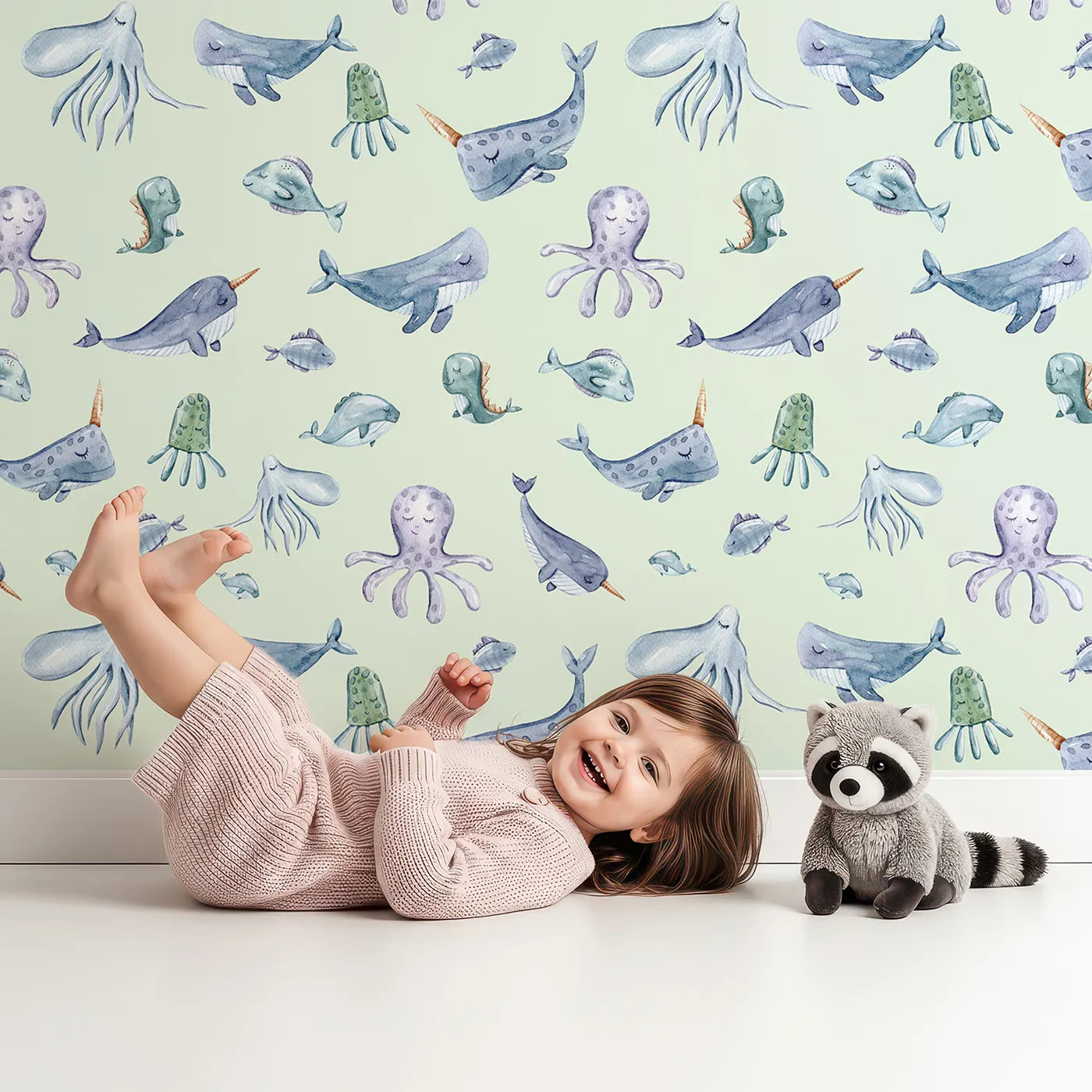 Lovely child standing in front of Whimsy Tots Gentle Ocean Parade Wallpaper in Mint Green with whale, narwhal, octopus, jellyfish, fish in design.