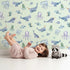 Lovely child standing in front of Whimsy Tots Gentle Ocean Parade Wallpaper in Mint Green with whale, narwhal, octopus, jellyfish, fish in design.