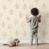 Lovely child standing in front of Whimsy Tots Floral Whisper Line Art Wallpaper in Peach with flower, leaf, blossom, lineart in design.