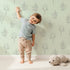 Lovely child standing in front of Whimsy Tots Floral Whisper Line Art Wallpaper in Mint Green with flower, leaf, blossom, lineart in design.