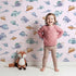 Lovely child standing in front of Whimsy Tots Dreamy Fish Parade Wallpaper in Pink with fish, fin, puffer, anglerfish, goby in design.