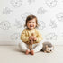 Lovely child standing in front of Whimsy Tots Line Art Camellia Wallpaper in White with flower, camellia, lineart, leaf in design.