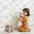 Lovely child standing in front of Whimsy Tots Line Art Bloom Wallpaper in White with flower, blossom, lineart, leaf in design.