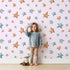 Lovely child standing in front of Whimsy Tots Starry Drift Wallpaper in Pink with star, watercolor, sky, night in design.