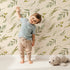 Lovely child standing in front of Whimsy Tots Botanical Breeze Wallpaper in Cream with leaf, sprig, branch, stem in design.