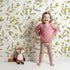 Lovely child standing in front of Whimsy Tots Golden Olive Garden Wallpaper in White with leaf, olive, stem, branch in design.