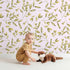 Lovely child standing in front of Whimsy Tots Golden Olive Garden Wallpaper in Blush with leaf, olive, sprig, stem in design.