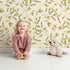 Lovely child standing in front of Whimsy Tots Golden Olive Garden Wallpaper in Cream with leaf, olive, stem, branch in design.