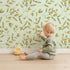 Lovely child standing in front of Whimsy Tots Golden Olive Garden Wallpaper in Mint with leaf, olive, sprig, branch in design.