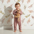 Lovely child standing in front of Whimsy Tots Sunset Meadow Breeze Wallpaper in White with leaf, branch, stem in design.