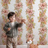 Lovely child standing in front of Whimsy Tots Vintage Garden Climb Wallpaper in White with flower, vine, bouquet, leaf in design.
