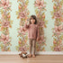 Lovely child standing in front of Whimsy Tots Vintage Garden Climb Wallpaper in Sage Green with flower, vine, leaf, bouquet in design.
