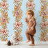 Lovely child standing in front of Whimsy Tots Vintage Garden Climb Wallpaper in Blue with flower, vine, leaf, bouquet in design.