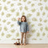 Lovely child standing in front of Whimsy Tots Gentle Palm Scatter Wallpaper in White with leaf, palm in design.