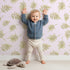 Lovely child standing in front of Whimsy Tots Gentle Palm Scatter Wallpaper in Blush Pink with leaf, palm in design.
