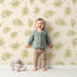 Lovely child standing in front of Whimsy Tots Gentle Palm Scatter Wallpaper in Cream with leaf, palm in design.