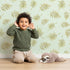 Lovely child standing in front of Whimsy Tots Gentle Palm Scatter Wallpaper in Sage Green with leaf, palm in design.