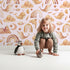 Lovely child standing in front of Whimsy Tots Whimsical Earth Friends Wallpaper in Blush Pink with whale, dolphin, narwhal, bird, fish, rainbow, moon, mountain, cloud in design.