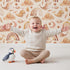 Lovely child standing in front of Whimsy Tots Whimsical Earth Friends Wallpaper in Cream with whale, dolphin, narwhal, bird, fish, rainbow, moon, mountain, cloud in design.