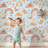 Lovely child standing in front of Whimsy Tots Whimsical Earth Friends Wallpaper in Sky Blue with whale, dolphin, narwhal, bird, fish, rainbow, moon, mountain, cloud in design.