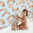 Lovely child standing in front of Whimsy Tots Sun-Drenched Shapes Wallpaper in Sky with rainbow, shape, brushstroke, line, dot in design.