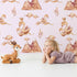 Lovely child standing in front of Whimsy Tots Feathered Serenity Wallpaper in Blush with bird, swan, crown, mountain, wing in design.