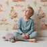 Lovely child standing in front of Whimsy Tots Feathered Serenity Wallpaper in Cream with bird, swan, crown, mountain, wing in design.