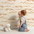 Lovely child standing in front of Whimsy Tots Soft Wave Rhythm Wallpaper in Sand with wave, curve, line, brushstroke, abstract in design.