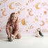 Lovely child standing in front of Whimsy Tots Sleepy Skies Shine Wallpaper in Blush Pink with moon, star, sky, sleepy face in design.
