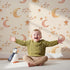 Lovely child standing in front of Whimsy Tots Sleepy Skies Shine Wallpaper in Cream with moon, star, sky, sleepy face in design.