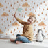 Lovely child standing in front of Whimsy Tots Rainy Day Nap Wallpaper in White with cloud, raindrop, sleepy face in design.
