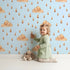 Lovely child standing in front of Whimsy Tots Rainy Day Nap Wallpaper in Blue with cloud, raindrop, sleepy face in design.