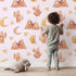 Lovely child standing in front of Whimsy Tots Desert Moon Dreams Wallpaper in Blush Pink with mountain, cactus, moon, star, sleepy face in design.