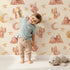 Lovely child standing in front of Whimsy Tots Desert Moon Dreams Wallpaper in Cream with mountain, cactus, moon, star, sleepy face in design.