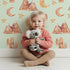 Lovely child standing in front of Whimsy Tots Desert Moon Dreams Wallpaper in Mint Green with mountain, cactus, moon, star, sleepy face in design.