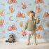 Lovely child standing in front of Whimsy Tots Desert Moon Dreams Wallpaper in Blue with mountain, cactus, moon, star, sleepy face in design.