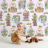 Lovely child standing in front of Whimsy Tots Whimsy Village Dreams Wallpaper in Blush Pink with house, rainbow, cup, jar, cloud, balloon in design.