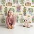 Lovely child standing in front of Whimsy Tots Whimsy Village Dreams Wallpaper in Beige with house, rainbow, cup, jar, cloud, balloon in design.