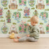 Lovely child standing in front of Whimsy Tots Whimsy Village Dreams Wallpaper in Mint Green with house, rainbow, cup, jar, cloud, balloon in design.