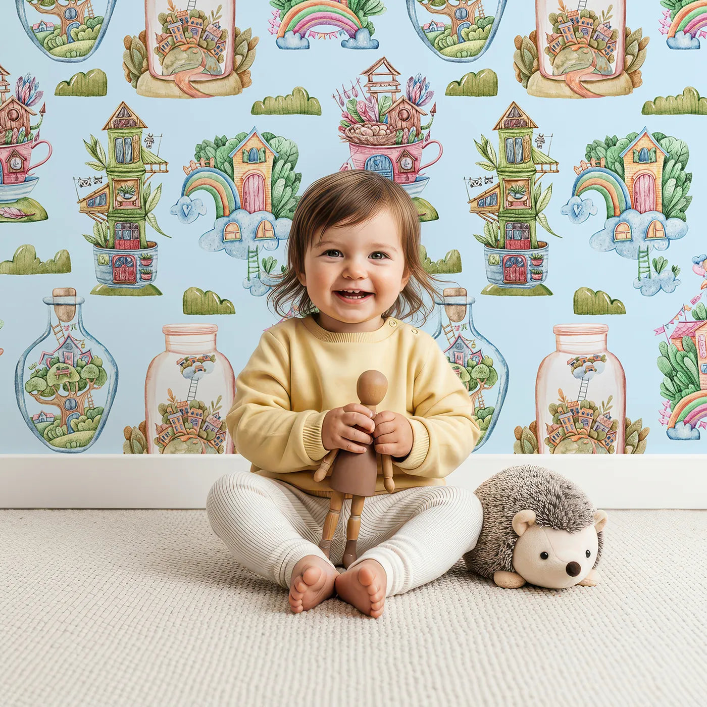 Lovely child standing in front of Whimsy Tots Whimsy Village Dreams Wallpaper in Blue with house, rainbow, cup, jar, cloud, balloon in design.