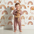 Lovely child standing in front of Whimsy Tots Golden Boho Rainbows Wallpaper in White with rainbow, star in design.