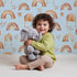 Lovely child standing in front of Whimsy Tots Golden Boho Rainbows Wallpaper in Blue with rainbow, star in design.